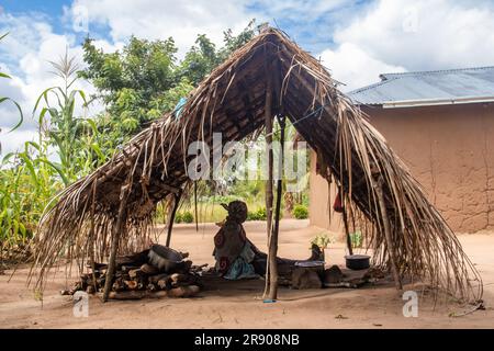 Eine alte Dame aus dem Makonde-Stamm sitzt in der primitiven Küche und bereitet lokale traditionelle Mahlzeit zu. Einfacher Metalltopf auf Feuer zwischen Steinen Stockfoto