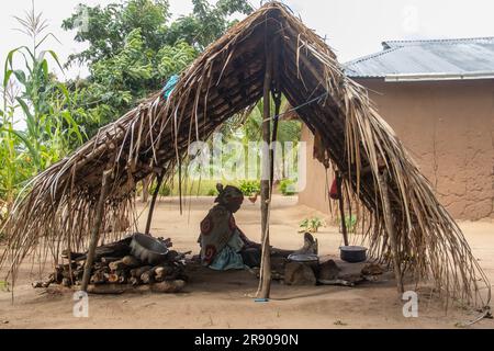 Eine alte Dame aus dem Makonde-Stamm sitzt in der primitiven Küche und bereitet lokale traditionelle Mahlzeit zu. Einfacher Metalltopf auf Feuer zwischen Steinen Stockfoto