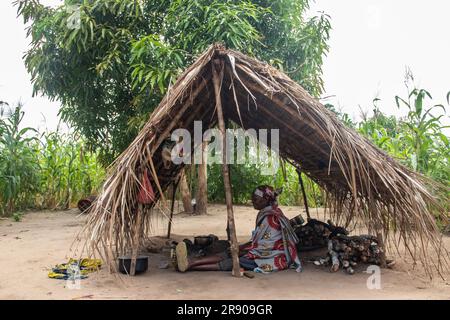 Eine alte Dame aus dem Makonde-Stamm sitzt in der primitiven Küche und bereitet lokale traditionelle Mahlzeit zu. Einfacher Metalltopf auf Feuer zwischen Steinen Stockfoto