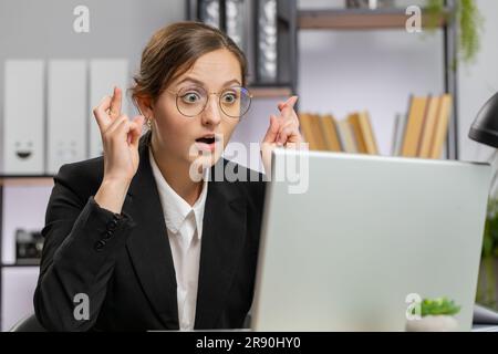 Eine überraschte Geschäftsfrau mit Brille drückte die Daumen, um auf den Laptop-Bildschirm zu schauen und nach Glücksnachrichten zu fragen. Sie wünschte gute Prüfungsergebnisse und träumte von einem Sieg im Lotto-Jackpot, einem Gewinn im Büro Stockfoto