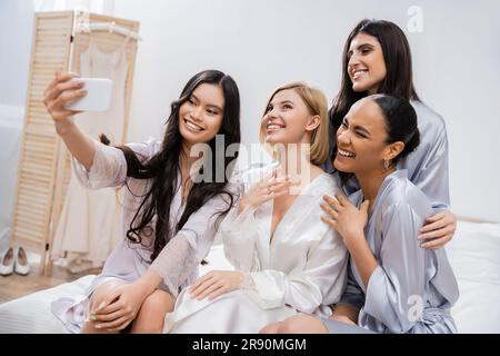 Vier Frauen, Brautparty, fröhliche blonde Braut und ihre gemischtrassigen Brautjungfern, die Selfie zusammen machen, Glück, Seidenroben, Verlobungsring, Brünette Stockfoto