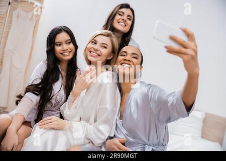 Vier Frauen, Brautparty, fröhliche blonde Braut und ihre gemischtrassigen Brautjungfern, die Selfie zusammen machen, Glück, Seidenroben, Verlobungsring, Brünette Stockfoto