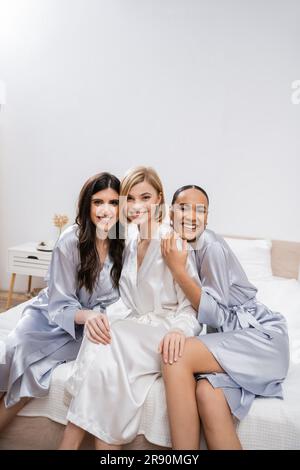 brautdusche, glückliche blonde Braut und ihre multikulturellen Brautjungfern sitzen auf dem Bett und zeigen Verlobungsring, Glück, Seidenmantel, Brünette und blond Stockfoto
