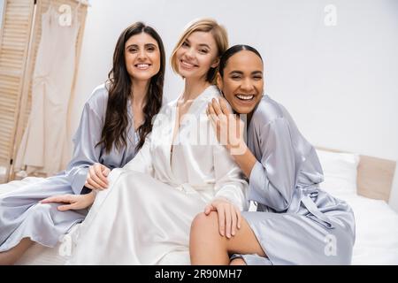 brautdusche, glückliche blonde Braut und ihre multikulturellen Brautjungfern sitzen auf dem Bett und zeigen Verlobungsring, Glück, Seidenmantel, Brünette und blond Stockfoto