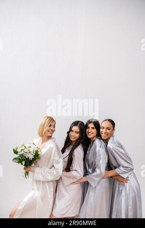 Positive Einstellung, fröhliche Braut mit Blumenstrauß und neben fröhlichen, gemischtrassigen Brautjungfern auf grauem Hintergrund, Rassenvielfalt, Seidenrobe Stockfoto