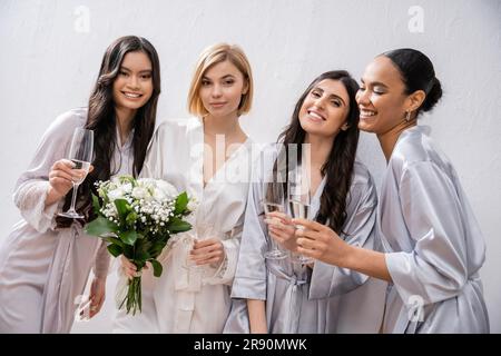 brautdusche, fröhliche multikulturelle Freundinnen, die Gläser mit Champagner halten, Braut mit weißen Blumen, brünette und blonde Frauen, Brautjungfern, Taucher Stockfoto