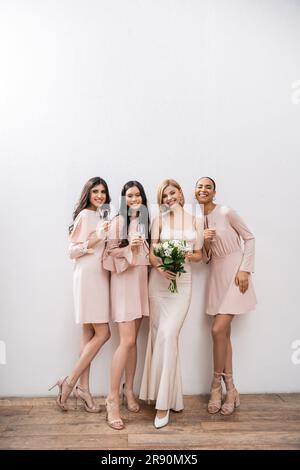 Positive Einstellung, blonde Braut im Hochzeitskleid mit einem Blumenstrauß und neben gemischtrassigen Brautjungfern mit Champagnergläsern auf grauem Hintergrund, rassisch Stockfoto