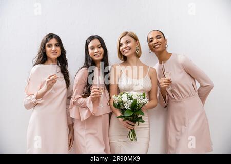 Positive Einstellung, glückliche Braut im Hochzeitskleid mit Brautstrauß und steht neben gemischtrassigen Brautjungfern auf grauem Hintergrund, Champagnergläser, Razzien Stockfoto