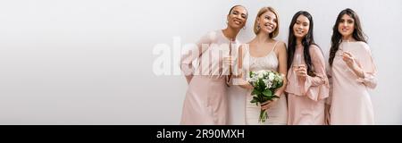 Positive Einstellung, glückliche Braut im Hochzeitskleid mit Brautstrauß und steht neben gemischtrassigen Brautjungfern auf grauem Hintergrund, Champagnergläser, Razzien Stockfoto