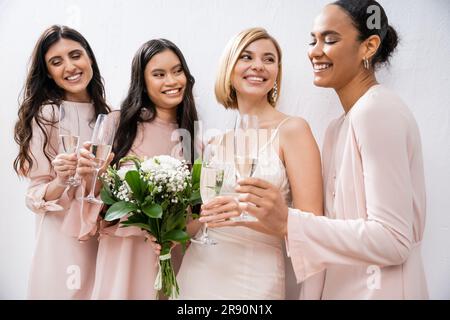 Positive Einstellung, blonde Braut im Hochzeitskleid, mit Brautjungfern, Champagnergläser, grauer Hintergrund, ethnische Vielfalt Stockfoto