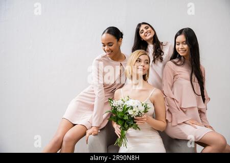 Hochzeitsfotografie, vier Frauen, fröhliche Braut und Brautjungfern, gemischtrassige Freundinnen, Hochzeitstag, kulturelle Vielfalt, auf einem Sessel sitzen, Graues Bac Stockfoto