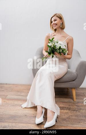 Hochzeitsfotos, besonderer Anlass, wunderschöne blonde Braut im Hochzeitskleid, sitzt im Sessel und hält ein Blumenstrauß auf grauem Hintergrund, weißer Fluss Stockfoto