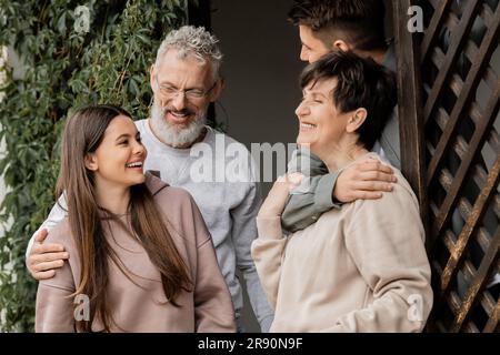 Lächelndes Teenager-Mädchen, das mit der Familie spricht, sich umarmt, während es den Elterntag feiert, und im juni zusammen auf der Veranda des Hauses steht, Eltern-Kind-Beziehung Stockfoto