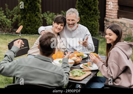 Lächelnde Eltern mittleren Alters, die sich während der grillparty in der Nähe von Sommeressen umarmen und mit Kindern sprechen, und Elterntagsfeiern im Garten im juni, schätzen Stockfoto