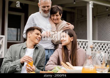 Lächelnde Kinder, die miteinander sprechen, während reife Eltern sich während der Elterntagsfeier im juni in der Nähe leckerer grillgerichte umarmen Stockfoto