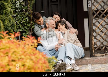 Lächelnde Kinder, die glückliche Eltern mittleren Alters umarmen, während sie zusammen auf der Veranda des Hauses sitzen und den Elterntag im Garten im juni feiern, Familie tr Stockfoto