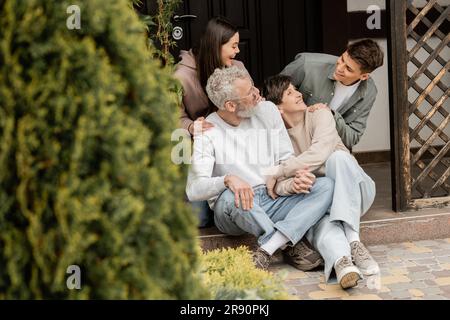 Fröhliche Kinder, die sich umarmen und mit Eltern mittleren Alters sprechen, während sie zusammen auf der Veranda des Hauses sitzen und den Elterntag im juni feiern, Familientradition Stockfoto