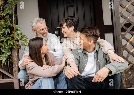 Positive Eltern mittleren Alters, die sich unterhalten, während sie Kinder umarmen und auf der Veranda des Hauses während der Elterntagsfeier im juni sitzen, Familientraditionen und Stockfoto