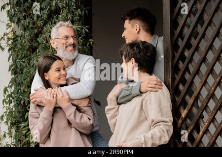 Positiver junger Mann, der Mutter mittleren Alters umarmt und mit Vater und Teenager Schwester während der Elterntagsfeier spricht, während er auf der Veranda des Hauses steht Stockfoto