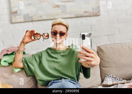 Zufriedene tätowierte Frau, die auf dem Sofa neben Kleidungsstücken sitzt und Selfie mit Sonnenbrille auf dem Smartphone macht, um online auszutauschen, nachhaltig zu leben und Stockfoto