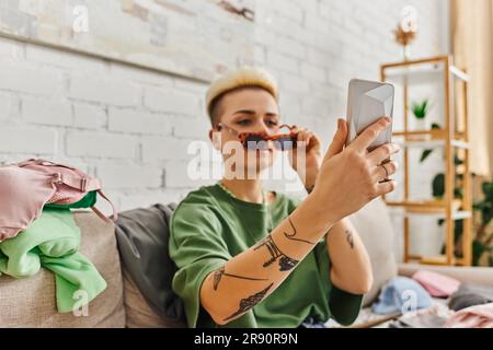 Tätowierte Frau mit trendigen Haaren nimmt Selfie in stilvoller Sonnenbrille für den Austausch auf dem Online-Marktplatz, persönliche Sachen werden enttarnt, nachhaltige Livi Stockfoto