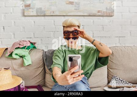 Junge und stilvolle tätowierte Frau mit Sonnenbrille, die Selfie auf dem Smartphone in der Nähe von Strohhut und Kleidung auf dem Sofa zu Hause macht, Online-Austausch, susta Stockfoto