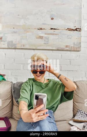 Junge, aufgeregte und tätowierte Frau, die Selfie in einer stilvollen Sonnenbrille auf einem Smartphone in der Nähe von Kleidung auf dem Sofa macht, um online in sozialen Medien zu tauschen, nachhaltig Stockfoto