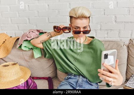 Stilvolle tätowierte Frau mit Sonnenbrille, die Selfie auf dem Smartphone macht, in der Nähe von Strohhüten und Kleidung auf dem Sofa, Online-Tausch, virtueller Marktplatz, nachhaltig Stockfoto