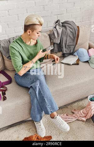 Sortieren von Kleidung, Online-Austausch, tätowierte Frau mit Smartphone, die ein Foto einer stilvollen Sonnenbrille macht, auf dem Sofa sitzt, nachhaltig lebt und achtsam ist Stockfoto