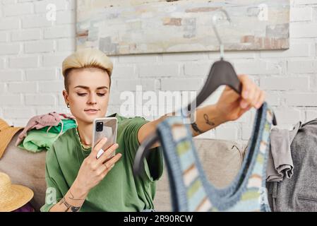 Virtueller Marktplatz, Online-Tausch, tätowierte Frau mit trendigen Haaren, die ein Foto von einem gestrickten Tanktop macht, auf dem Sofa neben Kleiderstücken sitzt, Sustain Stockfoto