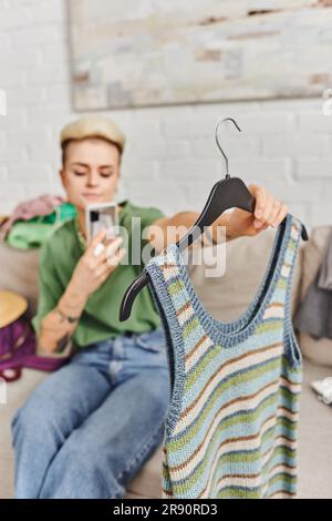 Junge, verschwommene Frau, die ein gestricktes Tanktop in der Hand hält und ein Foto mit dem Smartphone macht, um online auf dem Internet-Marktplatz zu tauschen, Kleidung zu entschlüsseln, zu halten Stockfoto