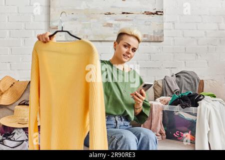 Online-Austausch, tätowierte Frau lächelt vor die Kamera, hält Smartphone und Kleiderbügel mit gelbem Pullover, sitzt auf dem Sofa neben Kleidung, nachhaltiges Leben Stockfoto