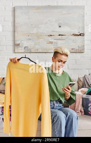 Junge tätowierte Frau mit gelbem Pullover und Blick auf Handy, während sie auf dem Sofa neben Kleiderschränken saß, Kleidung sortierte, online tauschte, susta Stockfoto