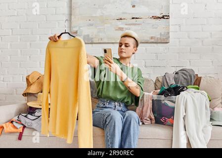 Tätowierte Frau mit trendigen Haaren, die auf dem Sofa neben Kleidung sitzt, wenn sie einen gelben Pullover für Online-Tausch auf dem virtuellen Marktplatz Sustaina fotografiert Stockfoto