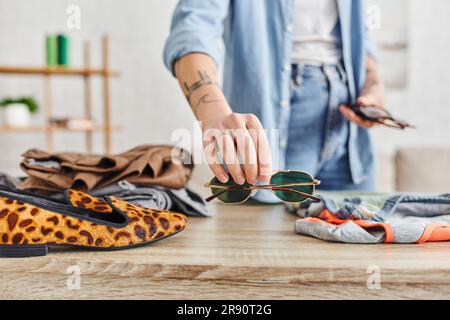 Zugeschnittener Blick auf eine tätowierte Frau, die eine Sonnenbrille in der Nähe des Tisches hält, mit Schuhen mit Tiermuster und gebrauchter Kleidung, dem Wiederverkaufsmarkt, nachhaltigem Wohnen und mi Stockfoto