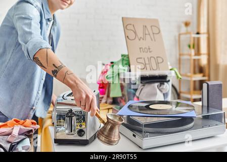 Teilansicht einer tätowierten Frau, die Cezve in der Nähe eines elektrischen Toasters, Schallplattenspielers, Kleidung und Karte mit Tausch hält, aber keine Ladenbeschriftung zu Hause, Sustai Stockfoto