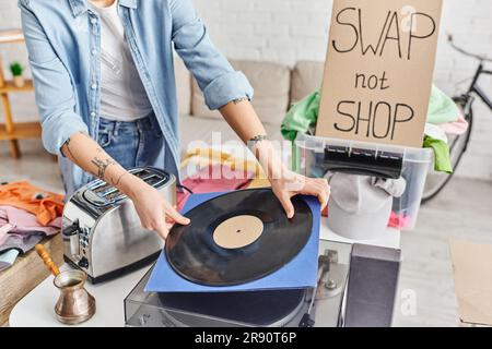 Zugeschnittener Blick auf eine tätowierte Frau, die eine Vinylscheibe in der Nähe eines Plattenspielers, eines elektrischen Toasters, einer Zezve, eines Plastikbehälters mit Kleidung und eines Tauschs, nicht einer Ladenkarte, hält. Stockfoto