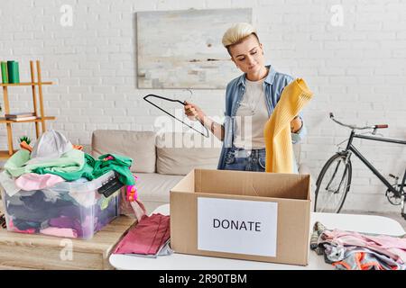 Tätowierte Frau mit trendigen Haarbügeln und gelbem Pullover in der Nähe eines Kunststoffbehälters mit Kleidung und Spendenbox im Wohnzimmer, nachhaltig Stockfoto