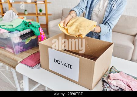 Teilansicht einer jungen tätowierten Frau, die einen gelben Pullover über einer Schachtel mit Spendenschrift neben einem Kunststoffbehälter mit Kleidung hält, nachhaltig Stockfoto