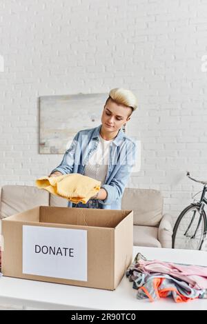 Wohltätige und ehrenamtliche Zwecke, junge tätowierte Frau mit gelbem Pullover in der Nähe von Kleidungsstücken und Karton mit Spendenschrift, nachhaltigem Wohnen und Stockfoto