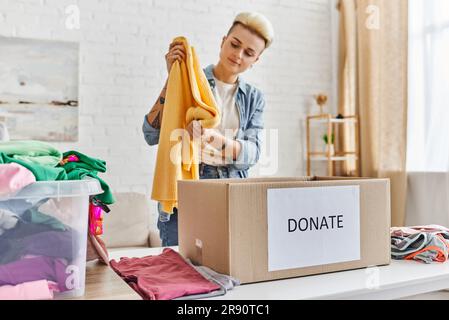 Junge und stilvolle Frau in legerer Kleidung mit gelbem Pullover neben einem Kunststoffbehälter mit Kleidung und Spendenbox im Wohnzimmer, nachhaltig Stockfoto