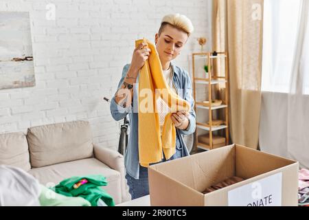 Ehrenamtliche und wohltätige, trendige und tätowierte Frau, die mit einem gelben Pullover neben der Kartonschachtel steht und Kleidung für Spenden zu Hause sortiert, nachhaltig Stockfoto