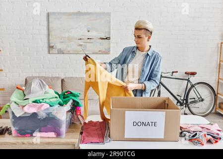 Lässig gestaltete und tätowierte Frau mit gelbem Pullover, während sie Kleidung in der Nähe eines Kunststoffbehälters und einer Kartonbox mit Spendenschrift sortierte, nachhaltig Stockfoto