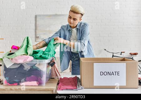 Förderung des sozialen Wohlergehens, junge tätowierte Frau, die Kleidung in einem Plastikbehälter neben einer Kartonschachtel sortiert, mit Spendenschrift im modernen Wohnzimmer, Sustain Stockfoto