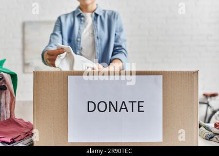 Gesellschaftliche Initiativen, partielle Sicht auf junge Frau, die Kleidung zu Hause in einer Schachtel mit Spendenschrift verpackt, verschwommener Hintergrund, nachhaltige Liv Stockfoto