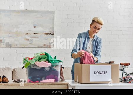 Spende für eine Sache, stilvolle tätowierte Frau, die Kleidung in Spendenbox neben Plastikbehälter mit Gebrauchtgegenständen im Wohnzimmer verpackt, nachhaltig Stockfoto