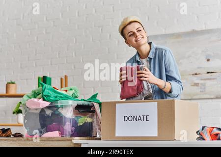 Fröhliche tätowierte Frau, die wegsieht, während sie mit Kleidung neben der Spendenbox und einem Plastikbehälter mit Kleidungsstücken zu Hause steht, nachhaltige Livi Stockfoto