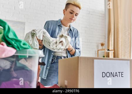 Spenden für eine Sache, lässig gestaltete und tätowierte Frau, die Kleidung in der Nähe von Plastikbehältern und Kartons mit Spendenschrift hält, nachhaltige Livi Stockfoto