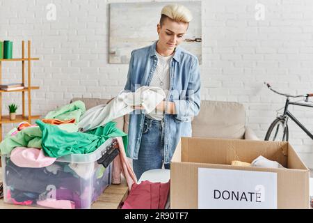 Tätowierte Frau mit trendigen Haaren, die auf eine Schachtel mit Spendenschrift schaut, während sie Garderobe im modernen Wohnzimmer sortiert, nachhaltiges Wohnen Stockfoto