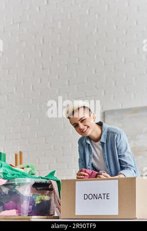 Junge und fröhliche Frau mit trendigen Haaren, die vorgeliebte Kleidung hält und in der Nähe von Plastikbehältern und Spendenboxen zuhause, Sustaina, auf die Kamera schaut Stockfoto